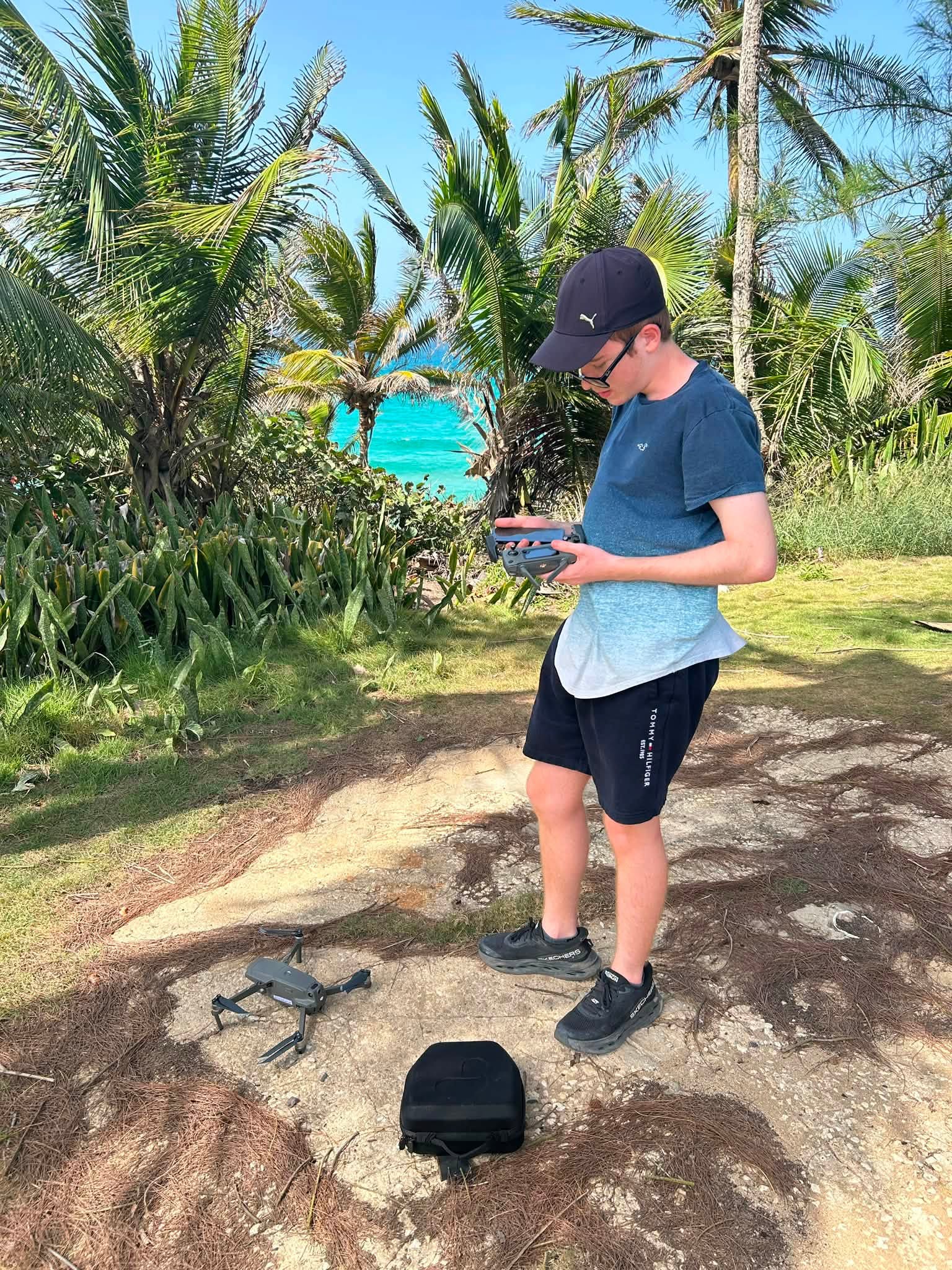 Drone operator holding a controller beside a drone, surrounded by palms
